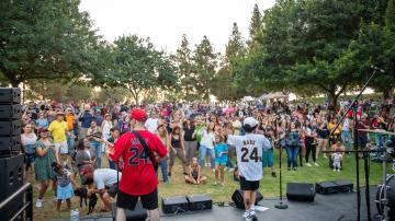 A band dressed in baseball jerseys on a stage plays to a large outdoor crowd surrounded by trees