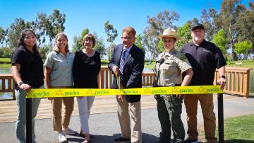 Six people, including OC Parks staff and Chairman Chaffee, stand behind a yellow ribbon. Chaffee prepares to cut the ribbon with large, ceremonial scissors.