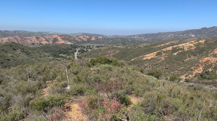 Hills covered with scrub with red cliffs in distance and blue sky above