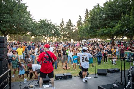 A band dressed in baseball jerseys on a stage plays to a large outdoor crowd surrounded by trees