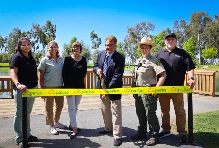 Six people, including OC Parks staff and Chairman Chaffee, stand behind a yellow ribbon. Chaffee prepares to cut the ribbon with large, ceremonial scissors.