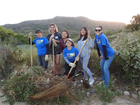 Six students with gardening tools stand surrounded by native plants with a pile of removed weeds in front of them.